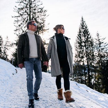 A man and a woman walking in the snow through a winter forest. It is a winter hike in Kleinwalsertal. | © Kleinwalsertal Tourismus eGen • Photographer: Oliver Farys