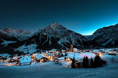 Winterliche Berglandschaft mit verschneiten Dörfern und Bergen unter einem klaren, dunklen Himmel. | © Kleinwalsertal Tourismus eGen • Fotograf: Robert Niederwolfsgruber