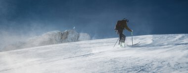 A skier in a winter mountain landscape, snowboarding down the slope amid strong wind and snowdrifts. | ©  Kleinwalsertal Tourismus eGen • Photographer: Robert Niederwolfsgruber