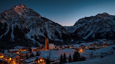 Winterliche Berglandschaft mit einem kleinen Dorf, das in der Nacht in warmem Licht erstrahlt, umgeben von schneebedeckten Bergen. | © Kleinwalsertal Tourismus eGen • Fotograf: Robert Niederwolfsgruber