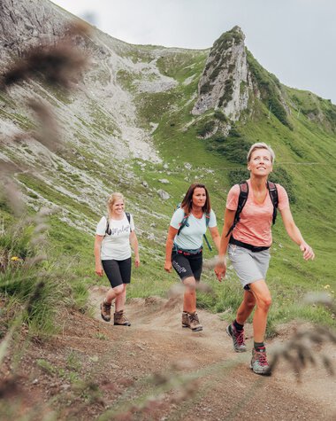 Three women hiking on a trail in a green mountain landscape with steep rocks in the background. | © Kleinwalsertal Tourismus eGen • Photographer: Stefan Klauser