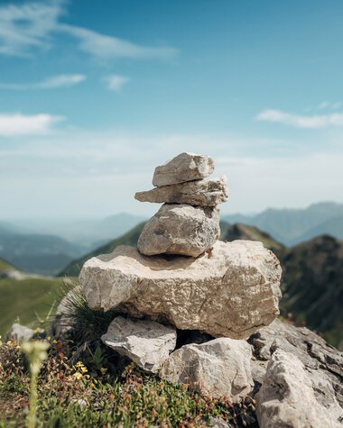 Cairn on a mountain summit with view of green hills and mountains in the background, blue sky with few clouds. | © Kleinwalsertal Tourismus eGen • Photographer: Stefan Klauser