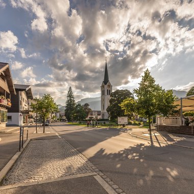 City view with church and sunlight shadows on the street under a cloudy sky in Riezlern. | © Kleinwalsertal Tourismus eGen • Photographer: Stefan Klauser