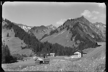 Historical black-and-white photo of a mountain village with several houses, surrounded by meadows and forests, with mountains in the background. | © Kleinwalsertal Tourismus eGen • Karl Max Kessler Archiv