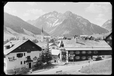 Black and white photo of a small Austrian village with mountains in the background, featuring a church, traditional houses, and cars on the street. | © Kleinwalsertal Tourismus eGen • Karl Max Kessler Archiv