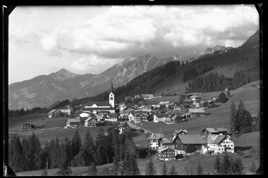 Black and white photo of the village Riezlern in the mountains, with many houses, a church in the center, and surrounding meadows and mountains. | © Kleinwalsertal Tourismus eGen • Karl Max Kessler Archiv