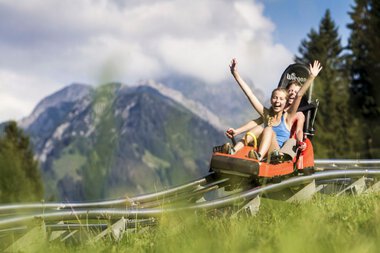 Two girls are laughing and cheering while riding a summer toboggan in the Alps, surrounded by green meadows and mountains. | © Kleinwalsertal Tourismus eGen • Oberstdorf Kleinwalsertal Bergbahnen • Photographer: Bastian Morell