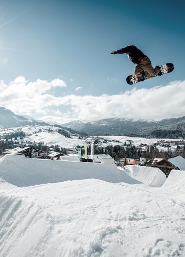 Snowboarder fliegt bei Sonnenlicht über eine Snowpark-Sprungrampe in einer verschneiten Winterszenerie. | © Kleinwalsertal Tourismus eGen • Crystal Ground • Fotograf: Stefan Eigner