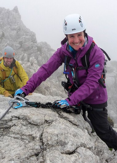 Two climbers on the Hindelanger Klettersteig, a smiling woman in a purple jacket, on a foggy rocky mountain wall. | © Kleinwalsertal Tourismus eGen • Alpinschule Oberstdorf