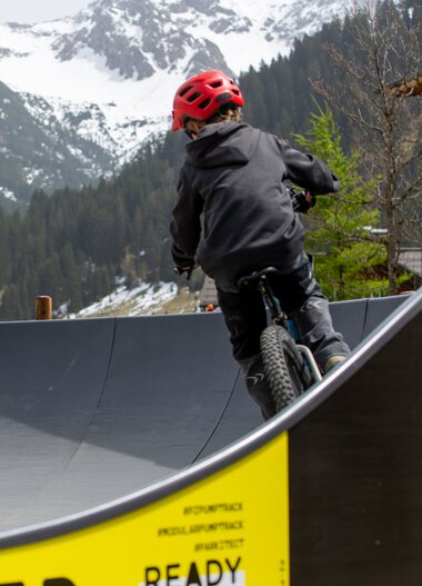 Two children riding bicycles on a pump track in a mountainous area with snow-capped peaks in the background. | © Kleinwalsertal Tourismus eGen • Photographer: Frank Drechsel