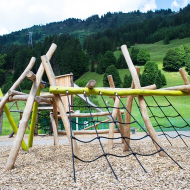 Playground with a wooden climbing frame and nets, surrounded by greenery, with mountains and trees in the background at Mittelberg. | © Kleinwalsertal Tourismus eGen