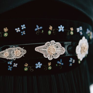 Close-up of traditional costume belt with delicate lace and pearl decorations, surrounded by floral embroidery in blue and green. | © Kleinwalsertal Tourismus eGen • Photographer: Stefan Klauser