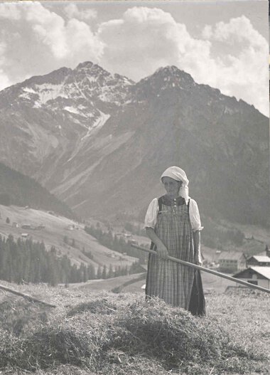 Historical black and white photo of two farmers in traditional attire raking hay in a field with mountain scenery in the background. | © Kleinwalsertal Tourismus eGen