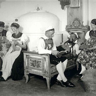 Historical black-and-white photo of five women in traditional costumes, sitting in a living room, engaged in handcrafts and playing music. | © Kleinwalsertal Tourismus eGen