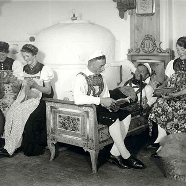 Historical black-and-white photo of five women in traditional costumes, sitting in a living room, engaged in handcrafts and playing music. | © Kleinwalsertal Tourismus eGen