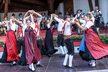 Dance group in traditional costumes performing at a folk festival in Kleinwalsertal, with musicians in the background. | © Kleinwalsertal Tourismus eGen • Photographer: Frank Drechsel