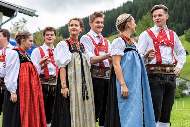 Group of people in traditional costumes at the Alphorn festival outdoors with mountains in the background. | © Kleinwalsertal Tourismus eGen • Photographer: Frank Drechsel
