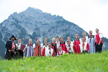 Group of people in traditional clothing in front of a mountain backdrop, some with musical instruments, standing on the grass. | ©  Kleinwalsertal Tourismus eGen • Photographer: Frank Drechsel
