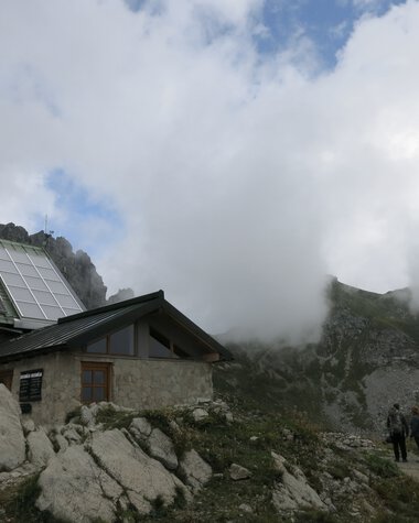 Mountain landscape with clouds, a small house, large rocks, and a group of people on a hiking trail | © Kleinwalsertal Tourismus eGen • Photographer: Antje Pabst