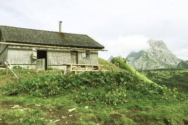 A rural house in the mountains with green meadows, mountains in the background, and cloudy sky. | © Kleinwalsertal Tourismus eGen