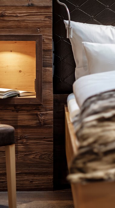 Innenansicht eines gemütlichen Zimmers mit einem Bett, Kopfteil in Schwarz, und einer Holzbox an der Wand, die ein Buch enthält. | © Bergchalet