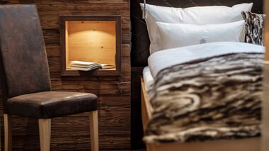 Interior of a cozy room with a bed featuring a black headboard, and a wooden wall box holding a book. | © Bergchalet