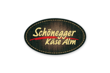 The logo of Schönegger Käse Alm, an oval-shaped design with a dark background and yellow, red, and white text. | © Schönegger Käsealm
