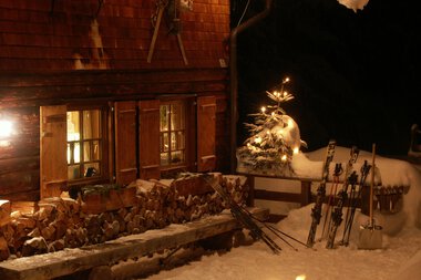Christmas hut at night with snow, lit windows, and ski equipment in the snow. Christmas decorations with string lights and a snowman. | © Kleinwalsertal Tourismus eGen