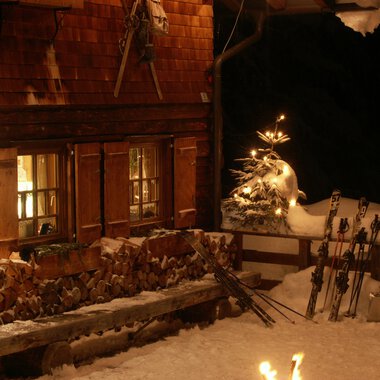 Die Weihnachtshütte bei Nacht mit Schneefall, beleuchtete Fenster und Skiern im Schnee. Weihnachtsdeko mit Lichterkette und Schneemann. | © Kleinwalsertal Tourismus eGen