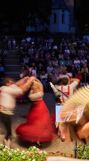 Musicians and dancers at the Alphorn Festival in Kleinwalsertal, with a large outdoor audience during dusk in the background. | © Kleinwalsertal Tourismus eGen • Photographer: Oliver Farys