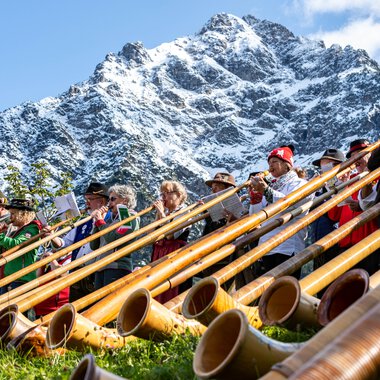 Musicians at the Alphorn Festival with a mountain backdrop, many alphorns lying on the grass. | © Kleinwalsertal Tourismus eGen • Photographer: Frank Drechsel