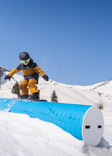 A child snowboarding in snowy mountains under a clear blue sky, riding on a curved blue rail. | © The Grilos • Chris Gollhofer